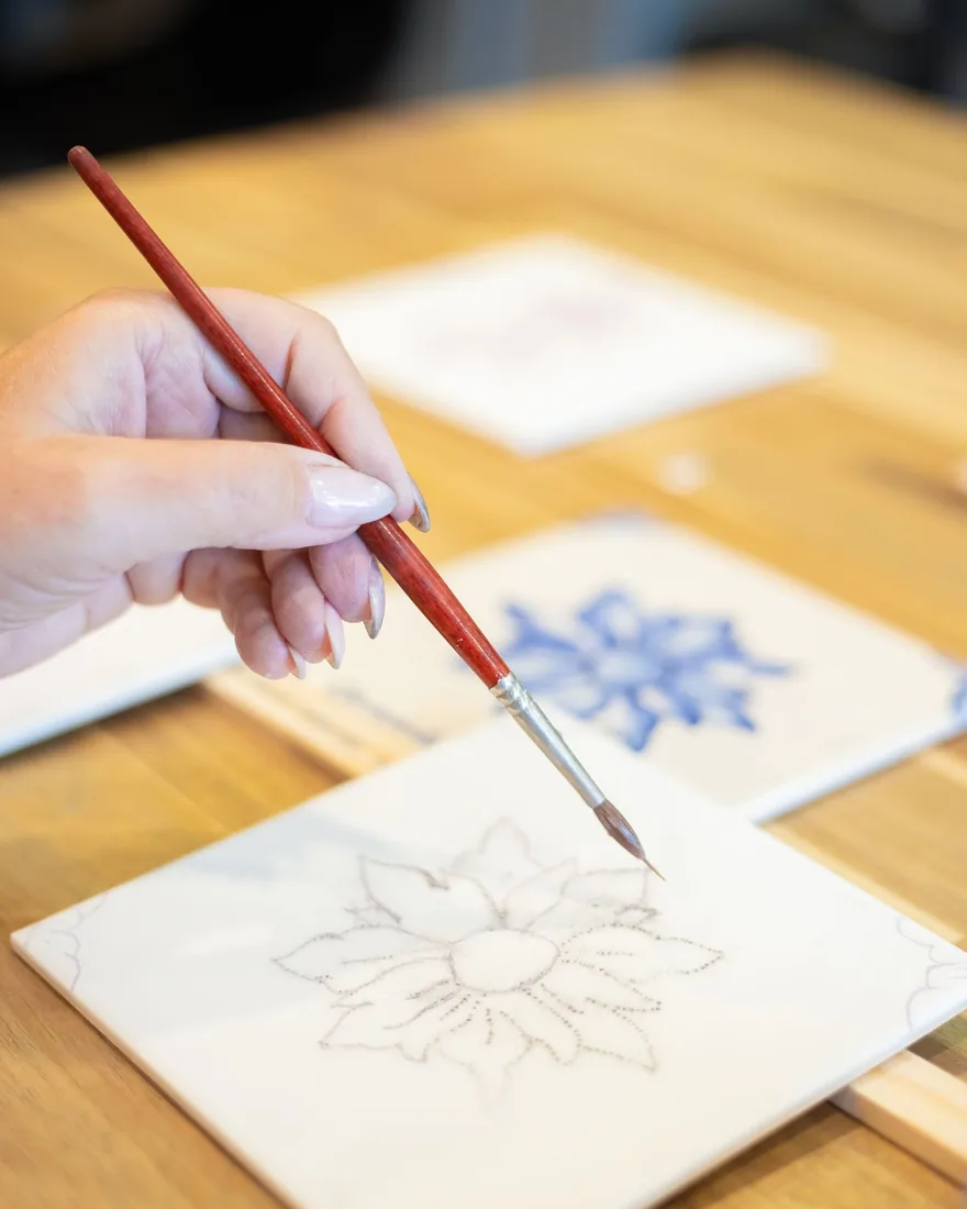 Mano sosteniendo un pincel fino sobre un azulejo blanco con un motivo floral transferido a puntos, junto a otras piezas pintadas en azul